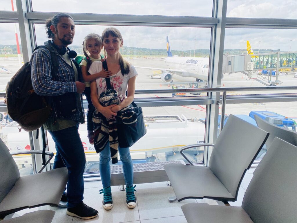 Una familia espera en la sala de embarque del aeropuerto con su equipaje. Al fondo, se ve un avión en la pista, listo para despegar. 