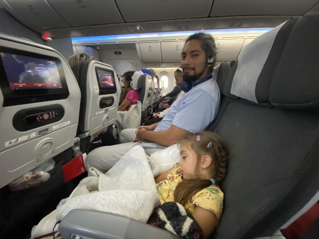 Un padre sonríe a la cámara mientras su hija duerme acurrucada y tapada con una manta en el avión. Ella disfruta de un sueño tranquilo durante el vuelo

